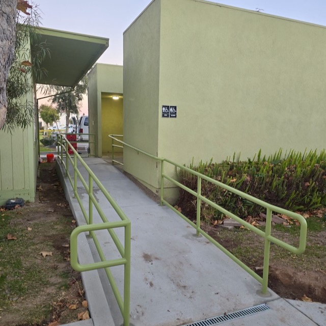 A completed ADA-compliant ramp featuring green metal railings installed along a concrete pathway, leading to a building with green walls.