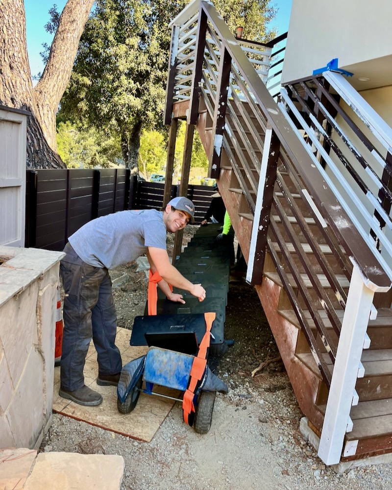 Team member carefully maneuvering the heavy steel staircase through a tight backyard using a dolly, showcasing the challenges of installation due to limited access at the San Luis Obispo project site.