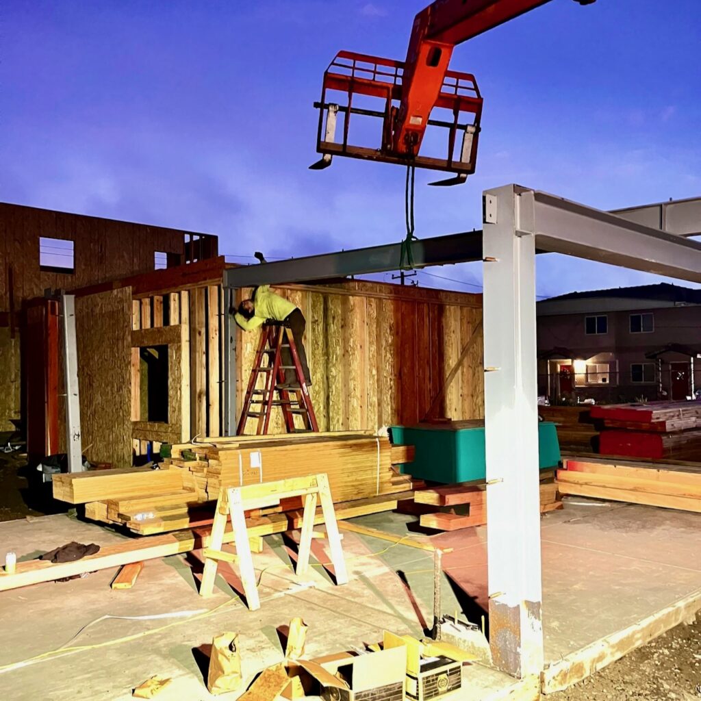 A worker installing structural steel framing at a construction site in Guadalupe, CA, as a crane lifts a steel beam into position during an evening installation.