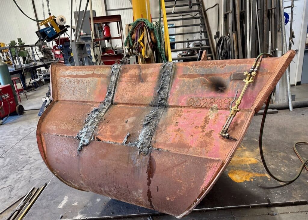 Excavator bucket during the repair process at J&M Welding Inc., showing worn-out pins and ears removed in preparation for new components to be installed.