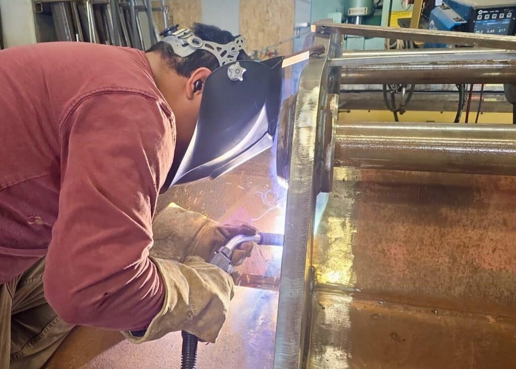 J&M Welding Inc. welder performing precision welding on an excavator bucket, ensuring accurate pin alignment for proper functionality in heavy-duty excavation tasks.