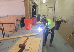 A J&M welder in protective gear works on the stainless steel countertops, welding and assembling components inside the shop. Tools and construction materials are visible on the covered surfaces, and precision welding ensures food-safe quality.